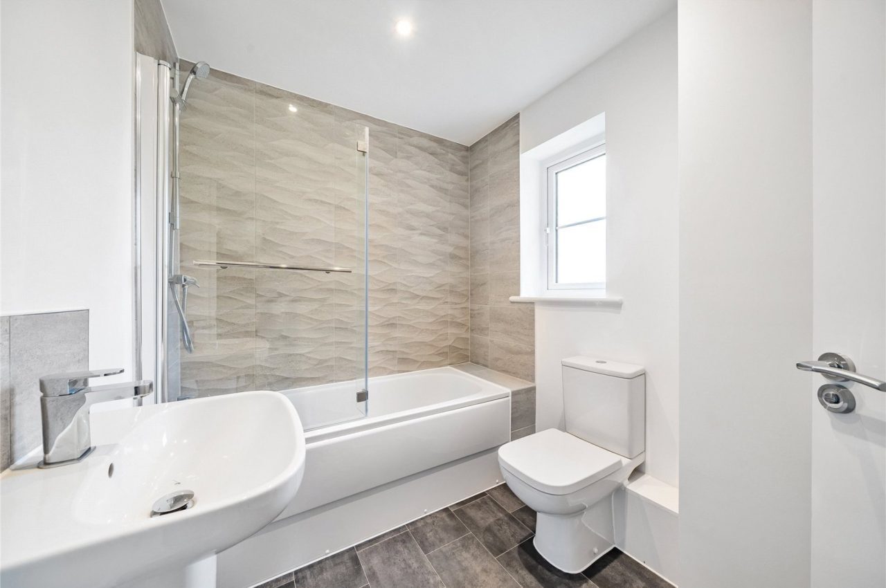 Bathroom with toilet, sink and bath tub, plus shower over the bath. Tiling is in a light grey. The bathroom also has a small window.